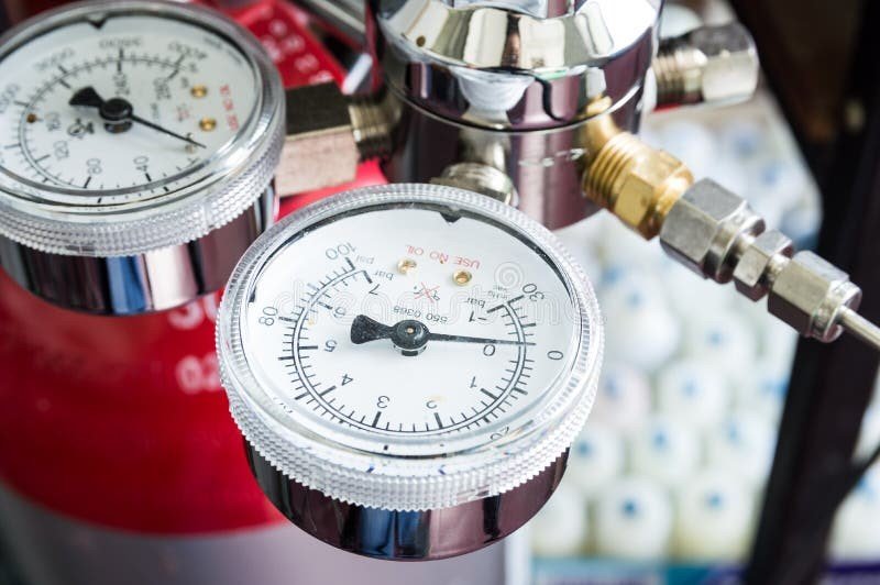 A close-up of a pressure gauge attached to a metallic valve system, with two round dials displaying measurements. The background includes a red cylinder and blurred white objects, suggesting a technical or industrial setting.