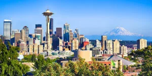 The image shows the Seattle skyline with the iconic Space Needle prominently featured among modern skyscrapers, set against a clear blue sky with Mount Rainier visible in the background.